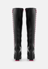 Botas de tacón negras hasta la rodilla con cremallera trasera y detalle de cordones rosas a lo largo de los lados, mostradas desde la parte trasera sobre un fondo blanco.