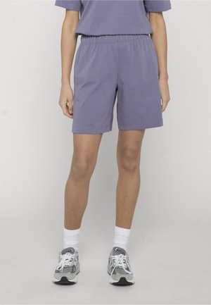 Osoba nosiaca fialové šortky s elastickým pásom, biele členkové ponožky a sivé bežecké topánky, stojaca pred jednoduchým svetlým pozadím.