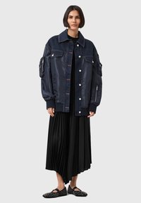 Una donna con capelli corti e scuri indossa un bomber blu navy oversize con tasche con zip, una gonna midi plissettata nera e scarpe Mary Jane con borchie nere.