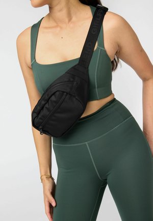 Ženska v zeleni športni modrček in pajkicah, ki nosi črno torbico z zadrgo in nastavljivim trakom čez ramo.