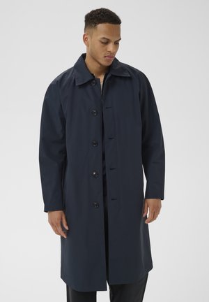 Man draagt een lange, donkerblauwe jas met knopen en een kraag, kijkt omlaag en staat tegen een effen lichtgrijze achtergrond.