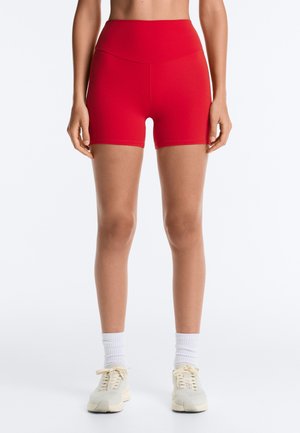 Vrouw draagt hooggetailleerde rode sportshort, witte crew-sokken en gebroken witte sneakers, staand tegen een effen witte achtergrond.