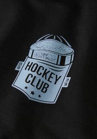 Tejido negro con un gráfico de un escudo azul claro desgastado que lleva el texto "HOCKEY CLUB" y el año "1978" junto con tres estrellas.