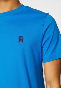 Blå bomuld t-shirt med rund halsudskæring. Har et lille broderet logo i rød og sort på venstre bryst. Korte ærmer.