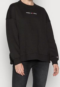 Persona che indossa un felpa nera di Tommy Jeans e jeans scuri, in piedi con le braccia rilassate ai lati contro uno sfondo semplice.