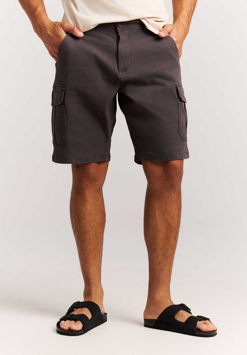 Homme portant un short cargo gris foncé avec des poches latérales, un t-shirt blanc et des sandales noires, debout les mains dans les poches.