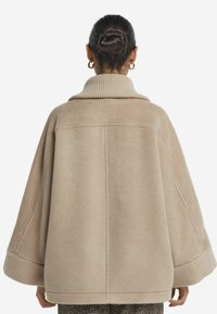 Beige ullkappa med hög ribbad krage, vida ärmar och textilstruktur. Har en lös passform och synliga sömnadsdetaljer på baksidan.