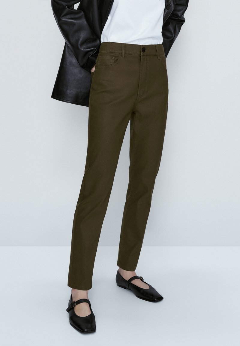 Personne portant un pantalon slim vert olive, une chemise blanche, une veste en cuir noire et des chaussures Mary Jane noires à bout carré, debout devant un fond blanc.