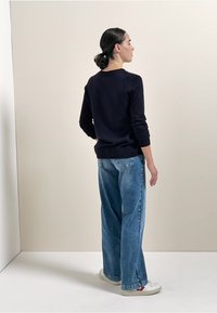 Marineblauer Pullover mit geripptem Saum, kombiniert mit hellblauen weit geschnittenen Jeans. Das Modell trägt weiße Sneaker mit roten Akzenten, von hinten betrachtet.