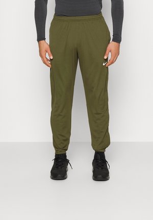 Mann trägt olivgrüne Nike-Sweathosen, schwarze Sneakers und ein langärmliges, dunkelgrau gestreiftes Oberteil, steht vor einem schlichten weißen Hintergrund.