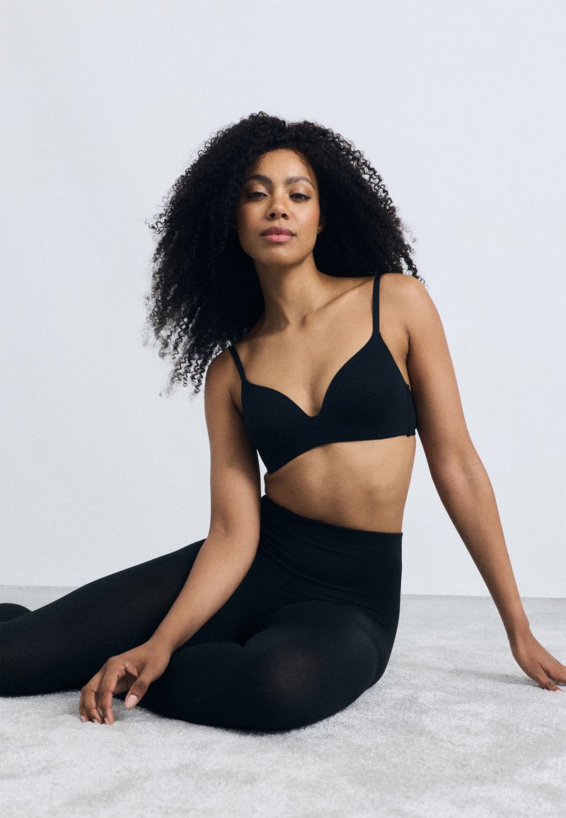 Schwarzer BH mit glatter Textur, V-Ausschnitt-Design, dünnen verstellbaren Trägern, kombiniert mit hoch taillierten schwarzen Leggings auf einer sanften grauen Oberfläche.