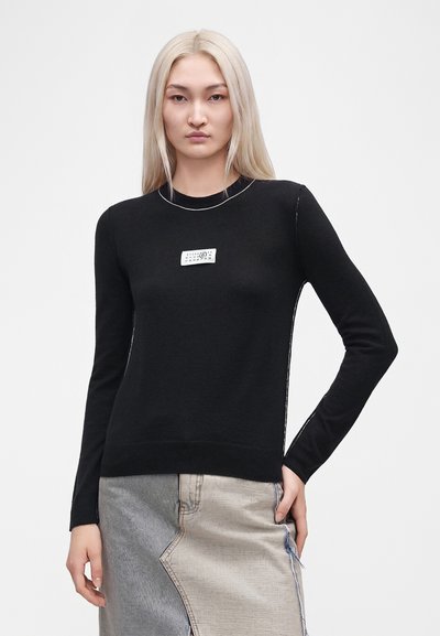 Femme aux longs cheveux blonds portant un haut noir à manches longues avec une étiquette blanche et une jupe en jean patchwork, debout avec une main sur la hanche.