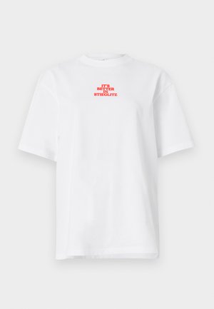 Young person wearing a white short-sleeve shirt with orange text, gray wide-leg trousers, black shoes, and a black leather cap, standing relaxed.