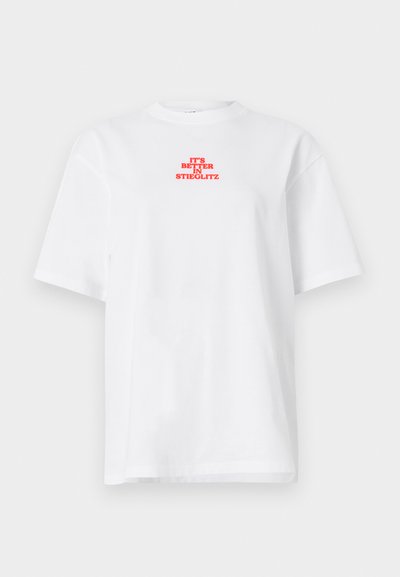 Λευκό κοντομάνικο μπλουζάκι με κόκκινο κείμενο "IT'S BETTER IN STIEGLITZ" κεντραρισμένο κοντά στο πάνω μέρος του στήθους.