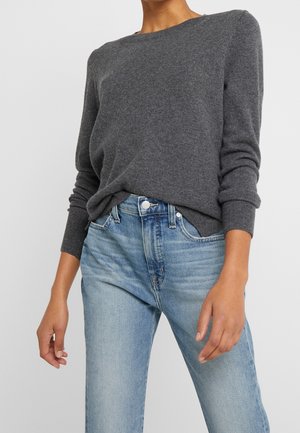 Person iført en langærmet, mørkegrå sweater, der er stoppet ned i lyseblå, højtaljede jeans mod en ensartet baggrund.