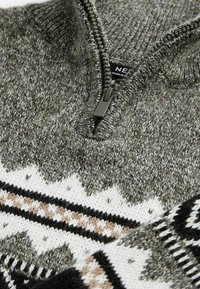 Gros plan d’un pull en maille gris avec une demi-fermeture éclair et des motifs géométriques blancs, noirs et marron sur la poitrine et les manches.