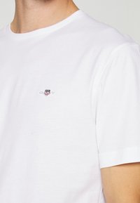 Nahaufnahme einer Person, die ein einfaches weißes T-Shirt mit einem kleinen Schild-Logo und dem Wort „GANT“ auf der linken Brust trägt.