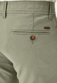 Helle grüne Hose aus einem glatten Stoff. Sie verfügt über eine Gesäßtasche mit einem Knopfverschluss und dezenten Steppnähten.