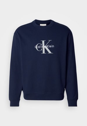 Tamno plavi pamučni dukserica s okruglim ovratnikom, dugim rukavima i rebrastim manžetama; sadrži veliki srebrni logotip s natpisima "Calvin Klein" i "CK."