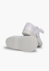 White mesh sneakers with perforated texture, flat rubber sole, and a soft fabric bow accent. Back view shows heel height and shape.