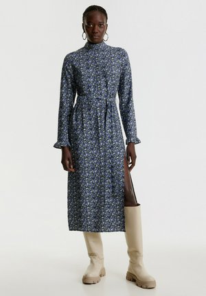 Blumenkleid in Midi-Länge mit blau-weißen Mustern, hohem Kragen, langen Ärmeln, tailliertem Schnitt und seitlichem Schlitz, kombiniert mit beigefarbenen Stiefeln bis zum Knie.
