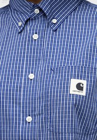 Μπλε πουκάμισο με λεπτές λευκές κάθετες ρίγες, λευκά κουμπιά, γιακά με κουμπιά και ταμπέλα Carhartt στην τσέπη στο στήθος.