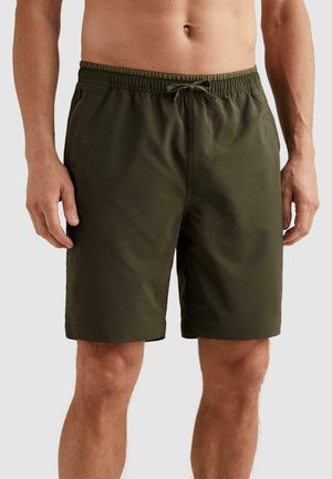 Muž nosiaci olivovo zelené šortky po kolená s elastickým pásom a šnúrkou, stojaci pred jednoduchým svetlým pozadím.