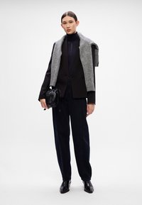 Čierny zúžený blazer s šedým pleteným šálom, tmavé džíny s vysokým pásom a čierne členkové topánky, drží malú čiernu kabelku. Hladké textúry.