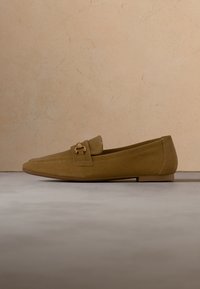 Un mocassin en daim marron avec un détail en métal doré sur le dessus, présenté sur un sol lisse devant un mur beige.