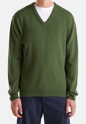 Mand iført en mørkegrøn V-hals sweater over en hvid T-shirt og mørkeblå bukser, der står mod en ensfarvet lys baggrund.