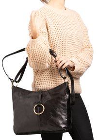 Borsa a mano in pelle nera con una texture liscia, caratterizzata da un accento circolare dorato e una zip superiore, indossata su una tracolla.
