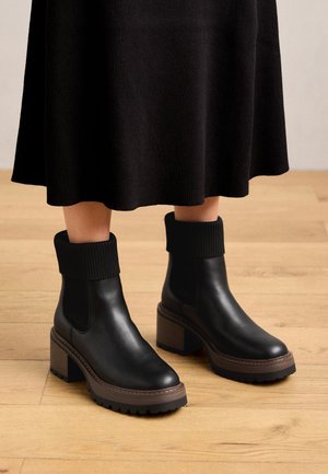 Bottes noires en cuir avec semelles épaisses marron et poignets en maille côtelée portées avec une jupe noire sur un sol en bois.