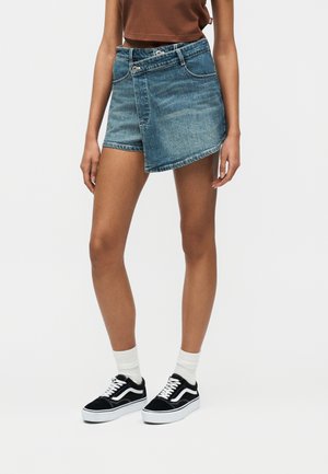 Γυναίκα που φοράει ασύμμετρο σορτς από τζιν, λευκές κάλτσες και μαύρα χαμηλά αθλητικά παπούτσια, στέκεται μπροστά σε ένα απλό λευκό φόντο.