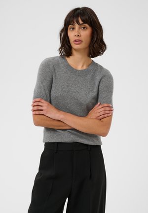 Vrouw met kort bruin haar, een grijze trui met korte mouwen en een zwarte broek, staand met gekruiste armen tegen een effen achtergrond.