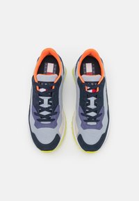 Scarpe sportive grigie e navy con dettagli arancioni, tomaia in rete testurizzata, sovrapposizioni viola e beige, e una suola gialla brillante.