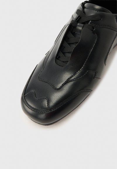 Μαύρο δερμάτινο αθλητικό παπούτσι με ραμμένα διακοσμητικά μοτίβα, μαύρα κορδόνια και λείο στρογγυλεμένο μπροστινό μέρος, απεικονίζεται σε ανοιχτό γκρι φόντο.