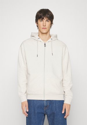 Cremefarvet zip-up hoodie med tekstureret mønster, snorhætte og ribbede manchetter. Båret med blå denim jeans.