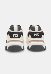 Par de zapatillas negras, blancas y beige con suelas gruesas vistas desde atrás, con el logo "NS" y una estrella en cada talón.