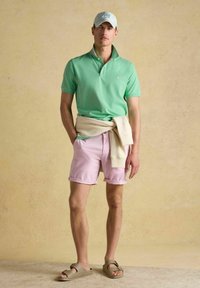 Zaļš polo krekls, rozā šorti un gaiša džemperis, sasiets ap jostasvietu. Beigžitu sandales. Fona ir teksturēta neitrāla krāsa.