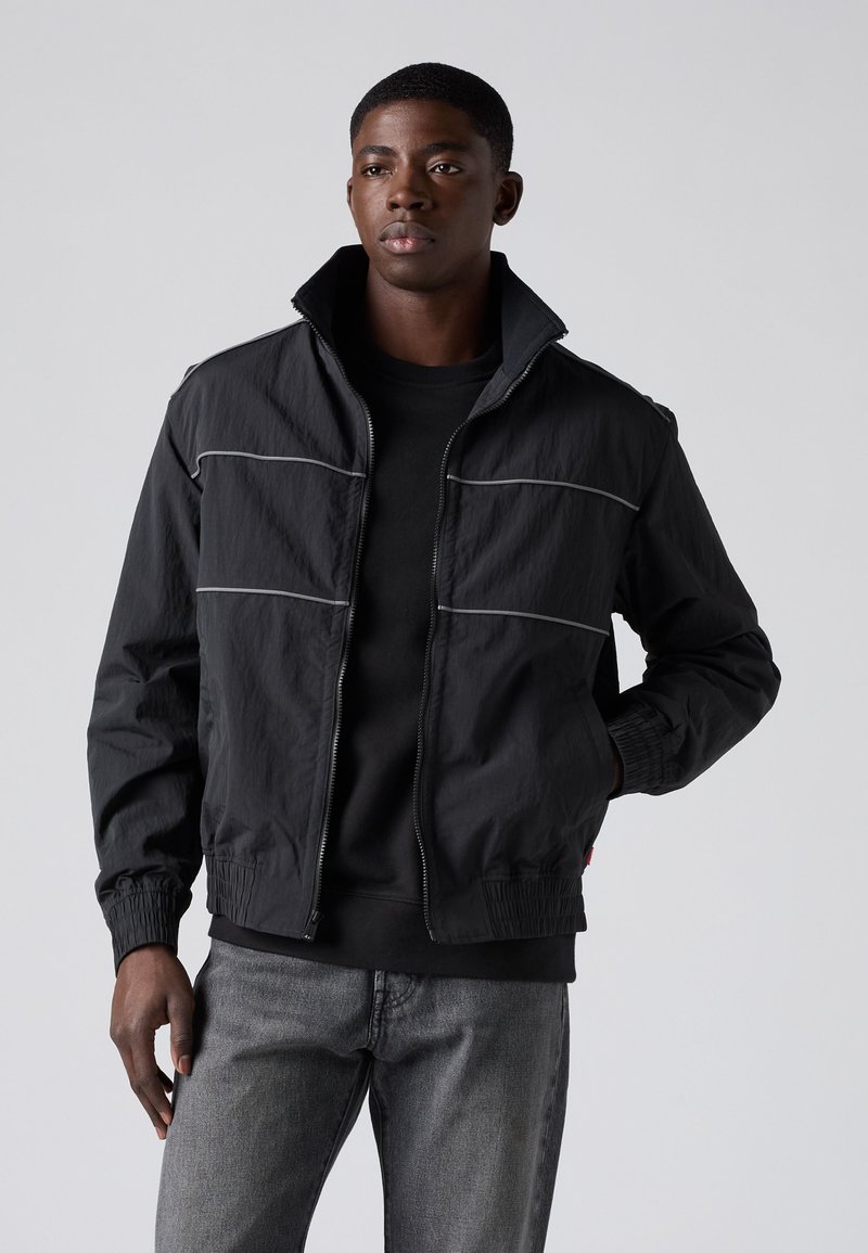 Jonge man met een zwart jack met ritssluiting en reflecterende biezen over een zwart shirt en grijze spijkerbroek, staand tegen een effen achtergrond.