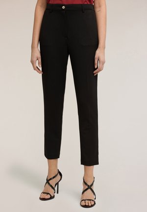 Femme portant un pantalon noir ajusté longueur cheville et des sandales à talons hauts à brides noires, debout devant un fond neutre.