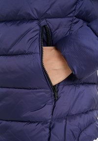 Giacca imbottita blu navy con tasca con zip, design trapuntato, texture liscia e vista ravvicinata di una mano che accede alla tasca.