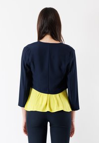 Giacca corta blu navy con peplum giallo. Maniche lunghe, scollatura rotonda e dettagli sulla cucitura posteriore. Tessuto liscio con colori a contrasto.