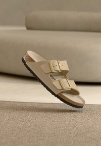 Μπεζ σανδάλι με διαφάνεια, δύο ρυθμιζόμενες χρυσές αγκράφες, μαλακό πέλμα και υφή μεσοφόρι από καφέ φελλό. Μαύρη λαστιχένια σόλα για καλύτερη πρόσφυση.