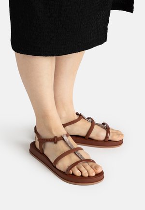 Füße, die braune flache Sandalen mit Riemen und verstellbaren Knöchelriemen tragen, und ein strukturiertes schwarzes Rock, das über den Knöcheln sichtbar ist.