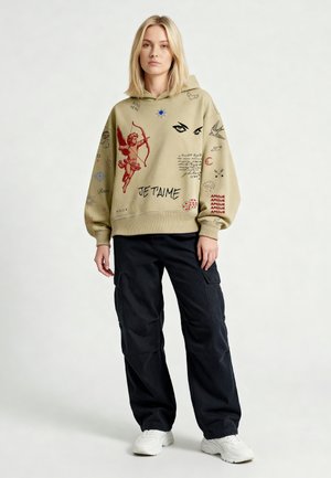 Junge Frau trägt einen beigen Hoodie mit Kupido-, Augen- und Textdrucken, kombiniert mit schwarzen Cargohosen und weißen Sneakers, steht vor weißem Hintergrund.