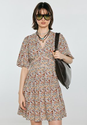 Kobieta ubrane w sukienkę w kwiatowy wzór, warstwowe naszyjniki, duże okulary przeciwsłoneczne i nosi ciemną, teksturowaną torbę na ramię.