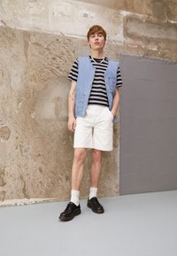 Ung mand iført en sort og grå stribet T-shirt, en lyseblå denimvest, hvide shorts, hvide sokker og sorte sko, står mod en tekstureret væg.