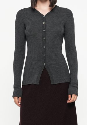 Vrouw die een aansluitend donkergrijs geribd vest met knopen en lange mouwen draagt, gecombineerd met een donkere rok.