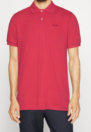 Homme portant un polo rouge à manches courtes avec deux boutons et un logo noir "GANT" sur le côté gauche de la poitrine, assorti à un pantalon foncé.